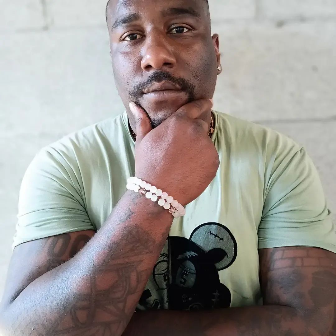 Close-up portrait of a tattooed man resting his hand under his chin, wearing a pale green graphic T-shirt and beaded bracelets.